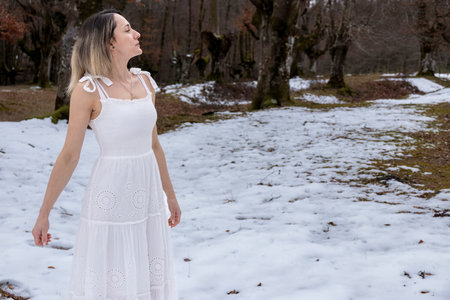a person in a white dress standing in a snowy forest, barefoot, with an overcast sky overheadの写真素材
