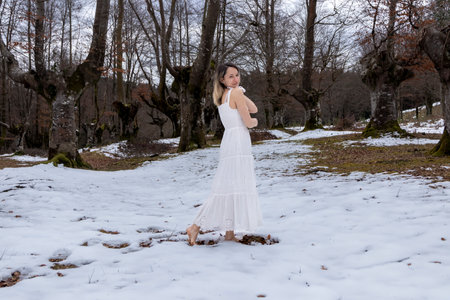 woman in a white dress walking in a snowy forest, barefoot, with an overcast sky overheadの写真素材
