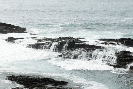 rocky coastline with light blue, rough waves crashing against black, jagged rocks under an overcast...