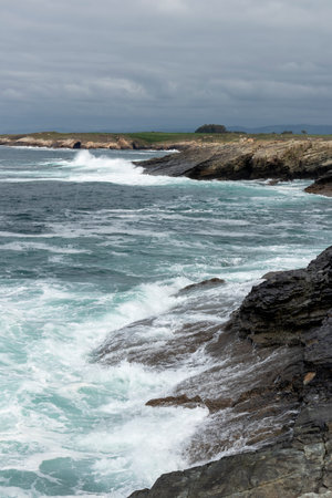 rocky coastline with turbulent waves crashing, under a cloudy sky, exuding a moody and dramatic atmosphereの写真素材
