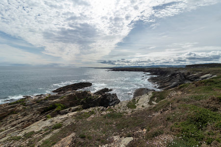 coastline with erosion, sparse vegetation, glistening ocean, and a partly cloudy sky. Serene and dynamic landscapeの写真素材