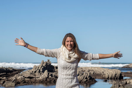 woman with arms outstretched stands by the rocky shore, facing the ocean waves under a clear skyの写真素材