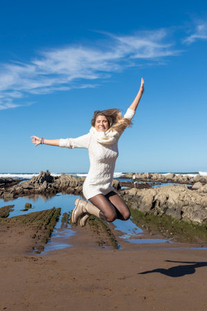 woman joyfully jumping on a beach, with rocky formations and a clear blue sky in the backgroundの写真素材