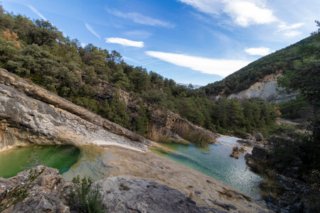 tranquil natural landscape with green water pools, rocky terrain, lush green trees, and a clear blue skyの写真素材