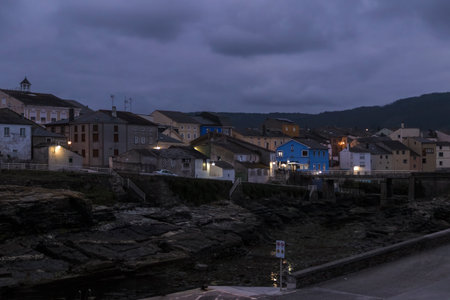 coastal town at dusk, with illuminated buildings nestled against a backdrop of darkening skies and rugged terrainの写真素材
