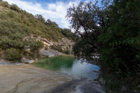 serene green pond nestled between rocky hills, surrounded by lush trees under a clear sky. It exudes natural beautyの写真素材