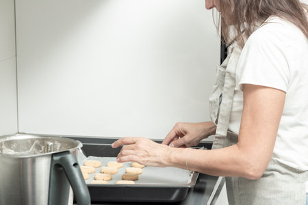 person arranging unbaked cookies on a tray beside a mixing bowl in a white kitchenの写真素材