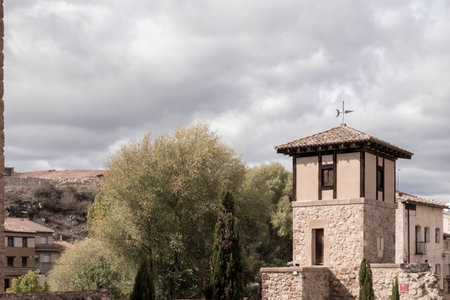 a stone building with a weathervane, surrounded by trees and structures, under a cloudy skyの写真素材