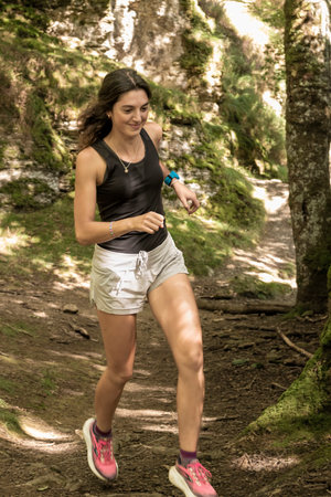 Smiling woman jogging on a forest trail surrounded by mossy rocks and trees during a sunny day.