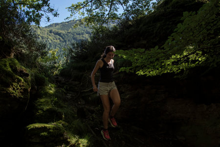 A woman hiking downhill through a shaded forest trail, surrounded by lush greenery, sunlight filtering through the trees, and a serene natural settingの写真素材