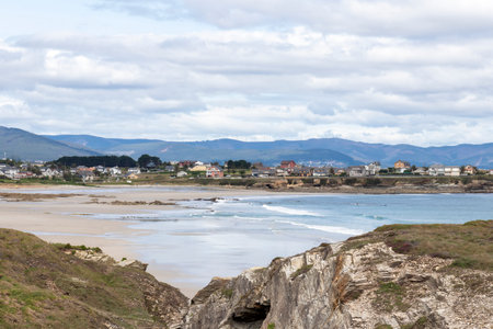 Coastal village with sandy beaches, rocky cliffs, and gentle waves under a cloudy sky, surrounded by rolling hills and scenic natureの写真素材