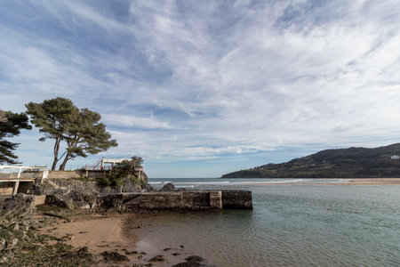 A serene coastal scene with a sandy beach, calm waters, and a small pier. A rocky cliff with trees...
