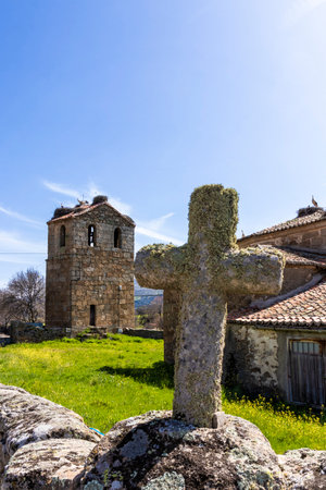 Mossy stone cross standing in front of an old church with stork nests on the roof on a sunny dayの写真素材