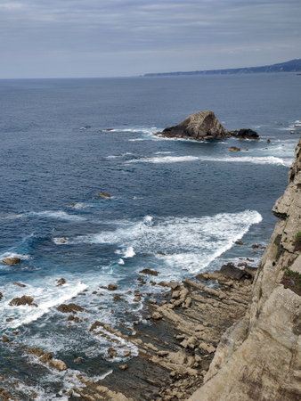 Breathtaking view of powerful ocean waves crashing against a rugged, rocky coastline, with a small island visible in the distance under a cloudy skyの写真素材