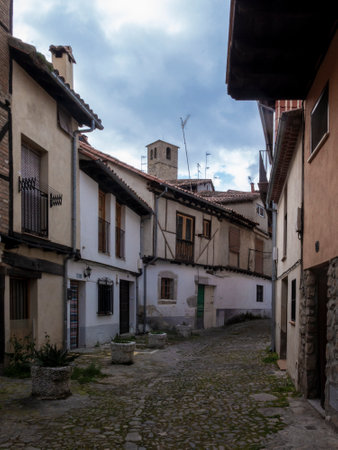 Narrow cobblestone street winding uphill between old houses with tiled roofs and a church tower in the background, in a spanish village under cloudy skyの写真素材