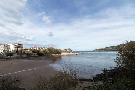 Tranquil coastal scene featuring a calm beach, ocean, buildings, and a cloudy skyの写真素材