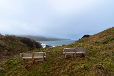 Scenic viewpoint with wooden benches offers breathtaking vista of rugged coastline, crashing waves, and misty hillsの写真素材