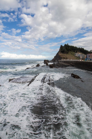 White ocean waves crashing over dark rocks near a hillside town under a blue cloudy skyの写真素材