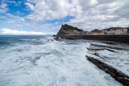 Foaming ocean waves hitting rocky formations on the coastline near a charming town under a cloudy skyの写真素材
