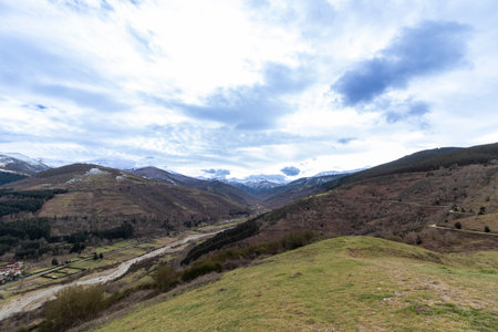 Stunning valley landscape showing snow-capped mountains, a winding river, and lush green hills under a cloudy skyの写真素材