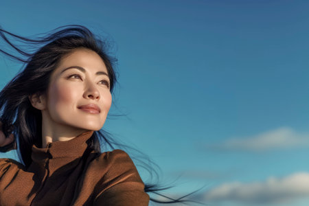 East asian woman looking up, hair blowing in wind against blue sky, symbolizing hope and freedomの素材
