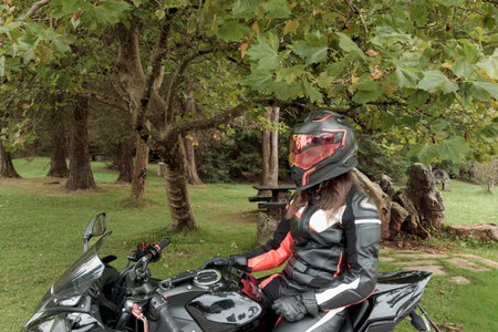 Woman motorcyclist wearing protective gear and helmet sitting on motorcycle in a green park