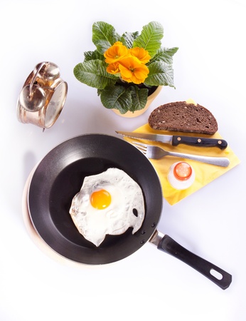 Fried egg in a frying pan with alarm-clock and yellow flowers on the white tableの写真素材