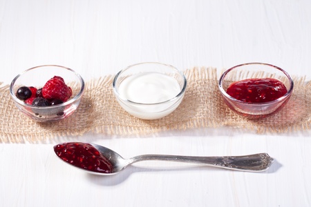 Bowls with fresh berries, jam and cream, served on white table with silver spoonの写真素材