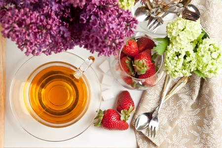 Top view on cup of tea and fresh strawberries served with lilac flowers and tableware