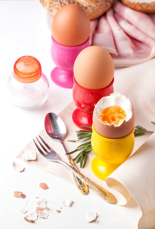 peeled boiled eggs in colorful cups on white table with salt in saltcellar, rosemary, silverware and bread with hamの写真素材