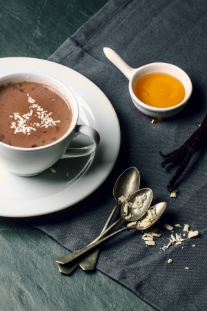 White cup of hot chocolate served with vintage spoons, honey and vanilla beans on dark gray napkinの写真素材
