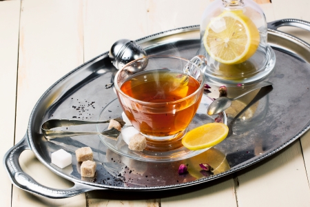 Glass cup of tea with sugar cubes, lemon and dry rose buds, served on silver vintage tray on white wooden table. See seriesの写真素材