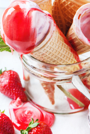 Close up of wafer cones with strawberry ice cream with syrup and fresh strawberries served in glass jar over white textile.の写真素材