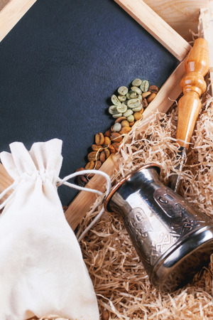 Green and brown decaf unroasted coffee beans with copper cezve and black empty chalkboard in wooden box with chips.の写真素材