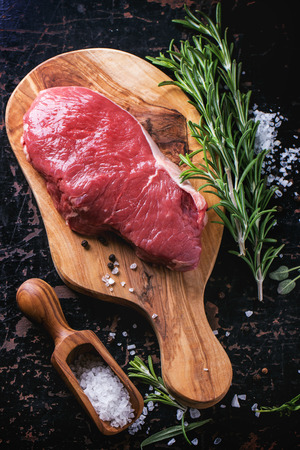 Raw steak on olive cutting board with rosemary herbs and red hot pepper over black wooden table. See seriesの写真素材
