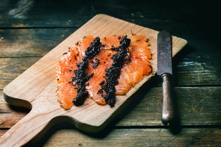 Pieces of salted salmon with black caviar on cutting board with vintage knife on old wooden table.の写真素材