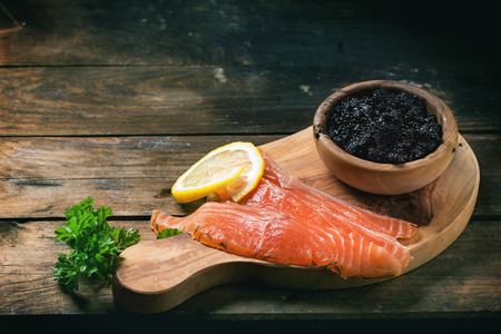 Bowl of black caviar and pieces of salted salmon on olive wood board with vintage knife over old wooden tableの写真素材
