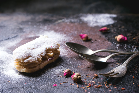 Eclairs with sugar powder, served with dry tea rose buds and vintage cutlery over dark background