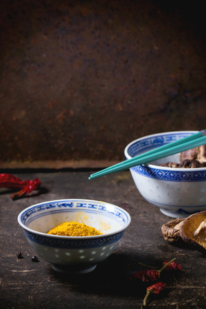 Tumeric powder and shiitake mushrooms in porcelain bowls, served with red hot chili peppers and turquoise chopsticks over dark tableの写真素材