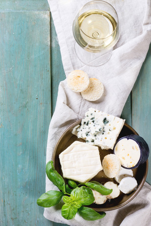 Variation of cheese, served with fresh basil, bread and glass of white wine over turquoise wooden table. Top view. See seriesの写真素材