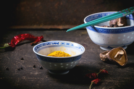 Tumeric powder and shiitake mushrooms in porcelain bowls, served with red hot chili peppers and turquoise chopsticks over dark table. See seriesの写真素材