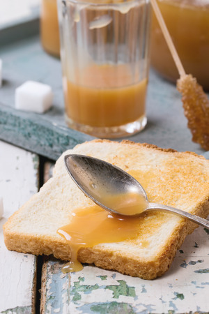 Toast with homemade caramel sauce for breakfast, served with spoon, jars and sugar cubes on gray tray over old wooden tableの写真素材