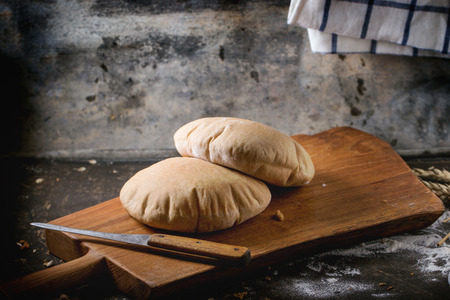 Two homemade wholegrain pita bread on wooden cutting board, served with knife and flour over dark tableの写真素材