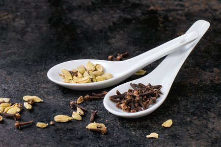 Cardamom seeds and cloves in white ceramic spoons over black backgroundの写真素材