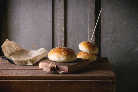 Fresh baked burger buns on small cutting board over wooden table. Dark rustic style.の写真素材