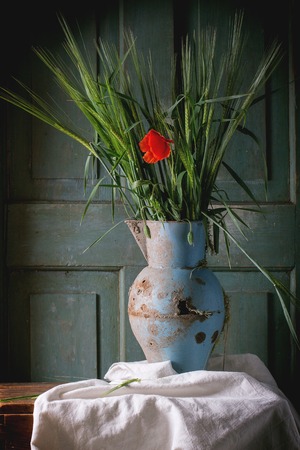 Bouquet of yang green barley with blooming red poppy flower in old metal vase on wooden table. Dark rustic atmosphereの写真素材