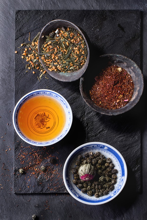 Assortment of dry tea and cup of hot tea. Green tea, black tae, green tea with rice, rooibos, dry rose buds in porcelan and old metal chinese bowls. Black textured slate background. Top view.の写真素材