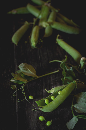 Heap of young green peas whole and broken with leaves and flower over old wooden table. Dark rustic style. Natural day light.の写真素材