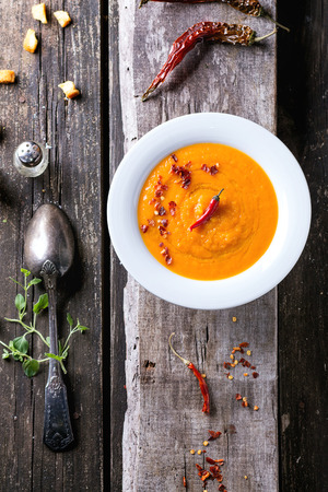 White plate of carrot cream soup with red hot chili pepper. Over old wooden table with vintage spoon, peppers, salt and croutons. Top view. Rustic style.の写真素材