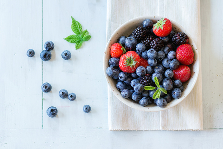 Spotted ceramic bowl with assortment berries blueberries, strawberries and blackberries at white textile napkin over wooden table. Natural day light. Top view